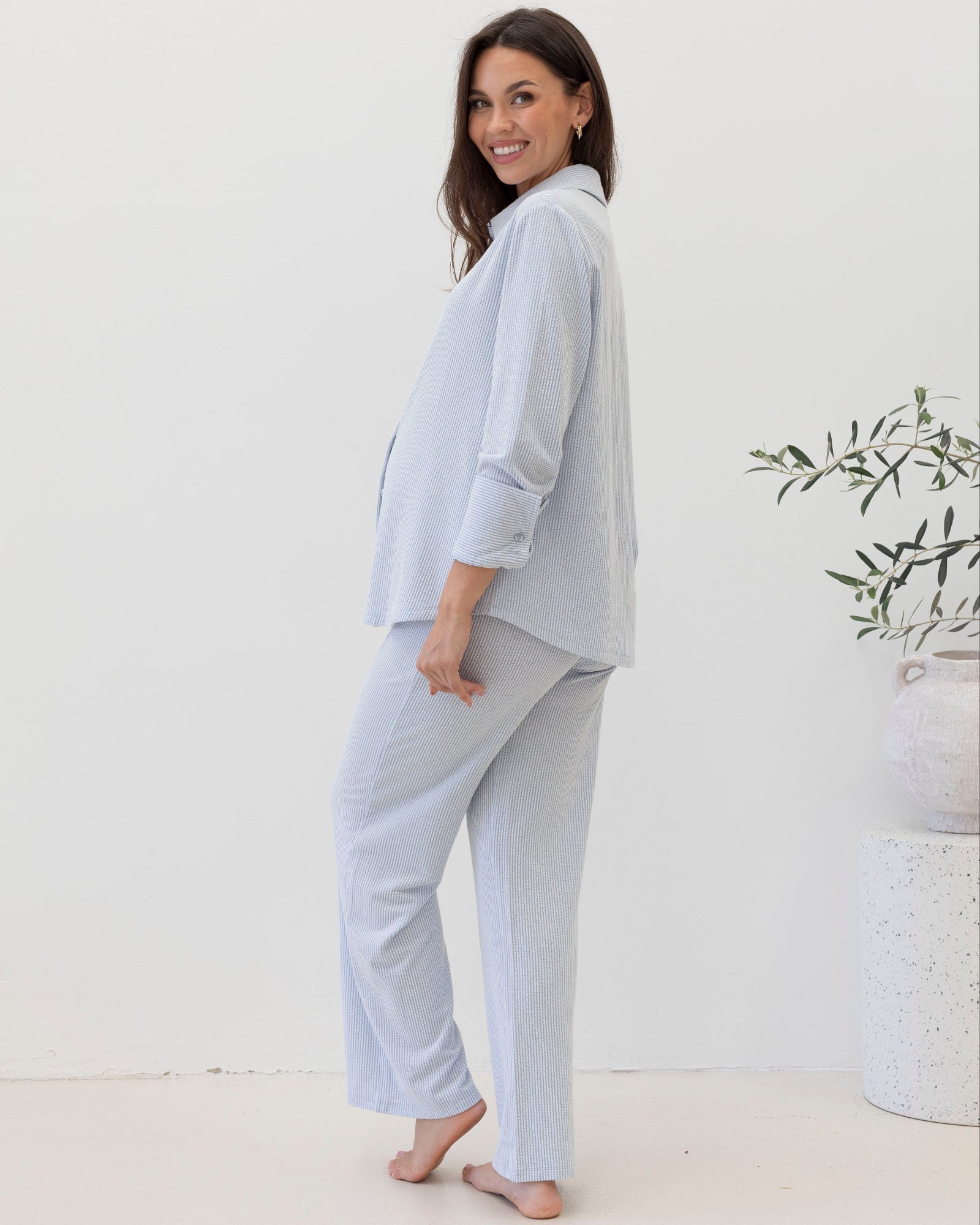 Woman wearing a light blue maternity pajama set standing in a minimalistic room with a plant.