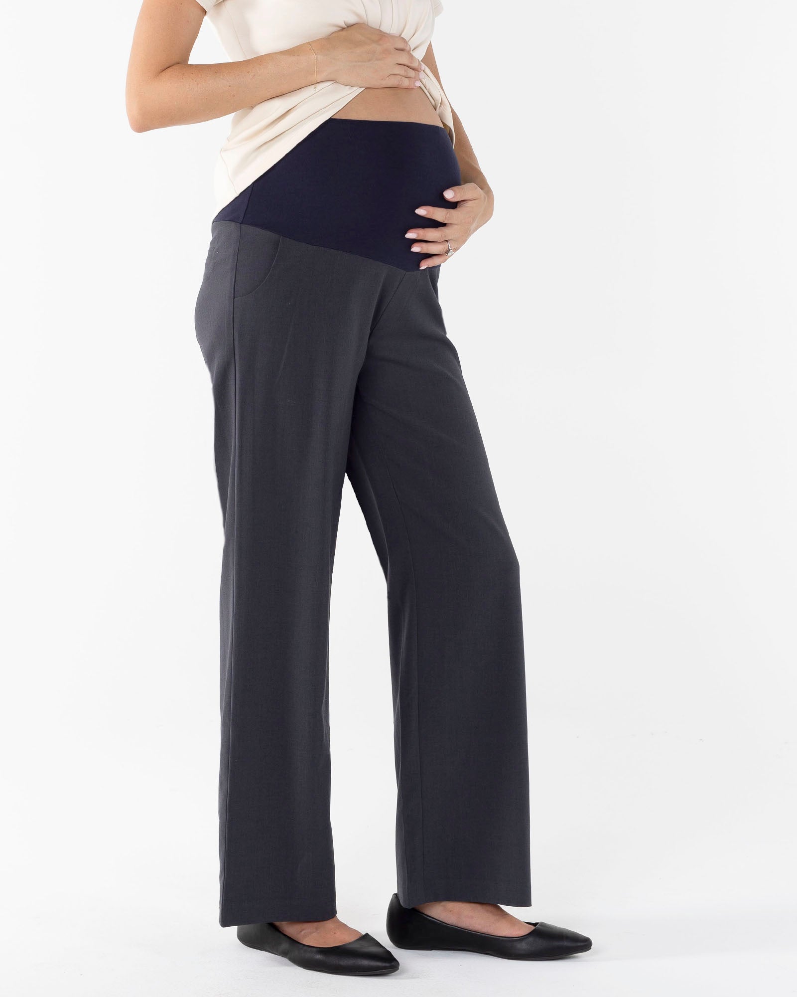 A pregnant woman wearing dark grey, relaxed fit maternity pants with an adjustable waistband. She is also wearing a white top and black shoes.