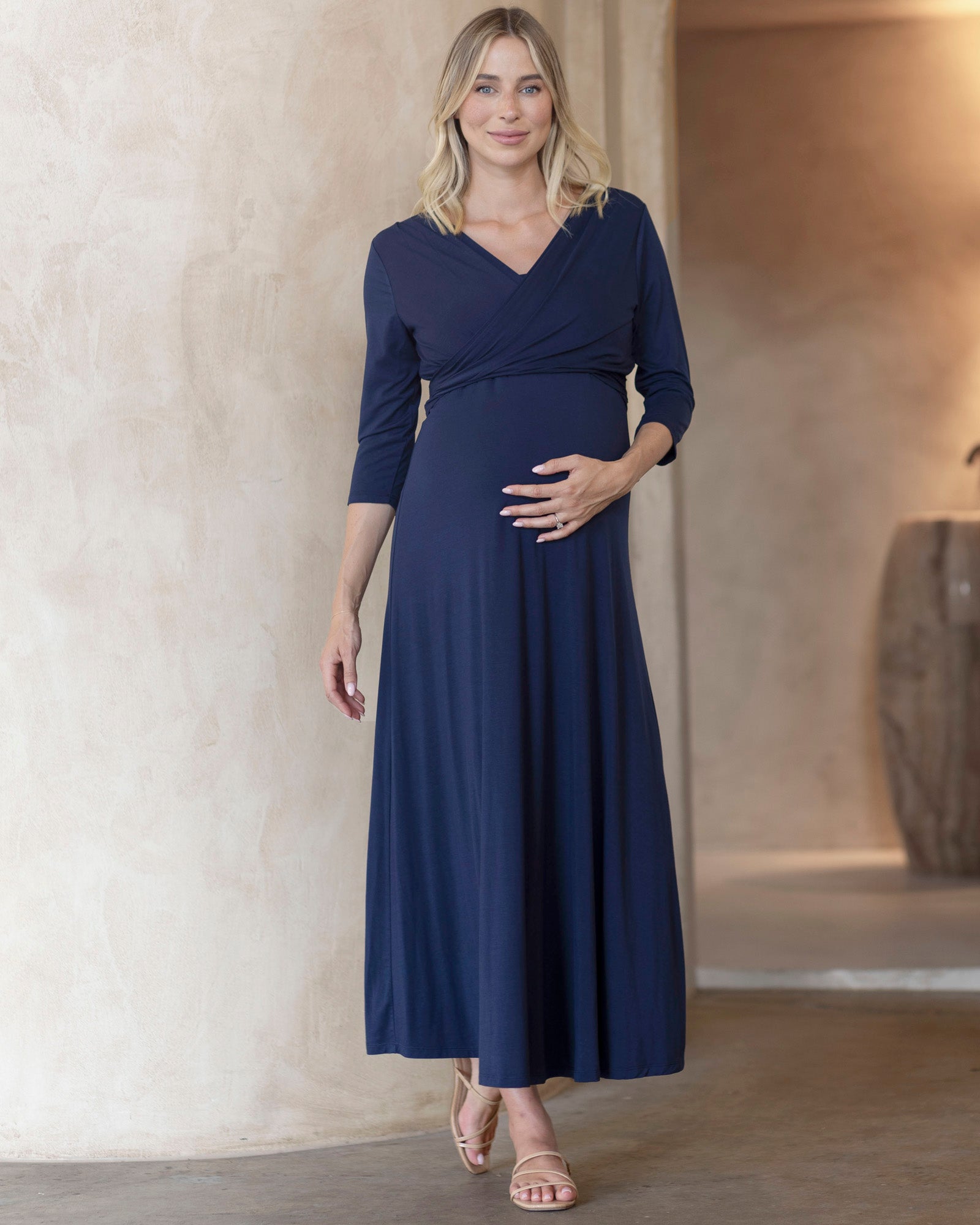 A pregnant woman wearing a navy-colored maternity maxi dress with a V-neckline and 3/4 sleeves, paired with a matching long sleeve wrap top.
