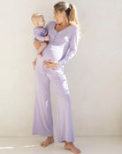 A pregnant woman wearing a lavender 3-piece maternity sleepwear set, holding a baby, with the top buttoned to accommodate a nursing need.