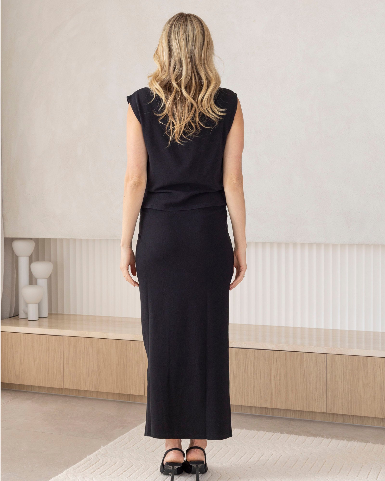 Woman wearing a black maternity maxi skirt standing in a minimalistic room with light-colored walls and wooden floor.