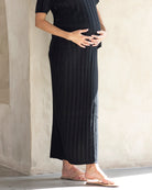 A pregnant woman wearing a wide rib knitted black maternity midi skirt with a straight-line silhouette, side seam split, and paired with a matching knit maternity top.