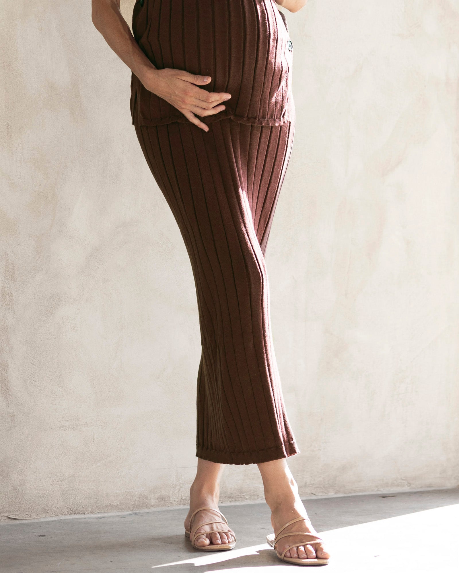 A pregnant woman wearing a brown, wide rib knitted maternity midi skirt with a side split, paired with a matching top.