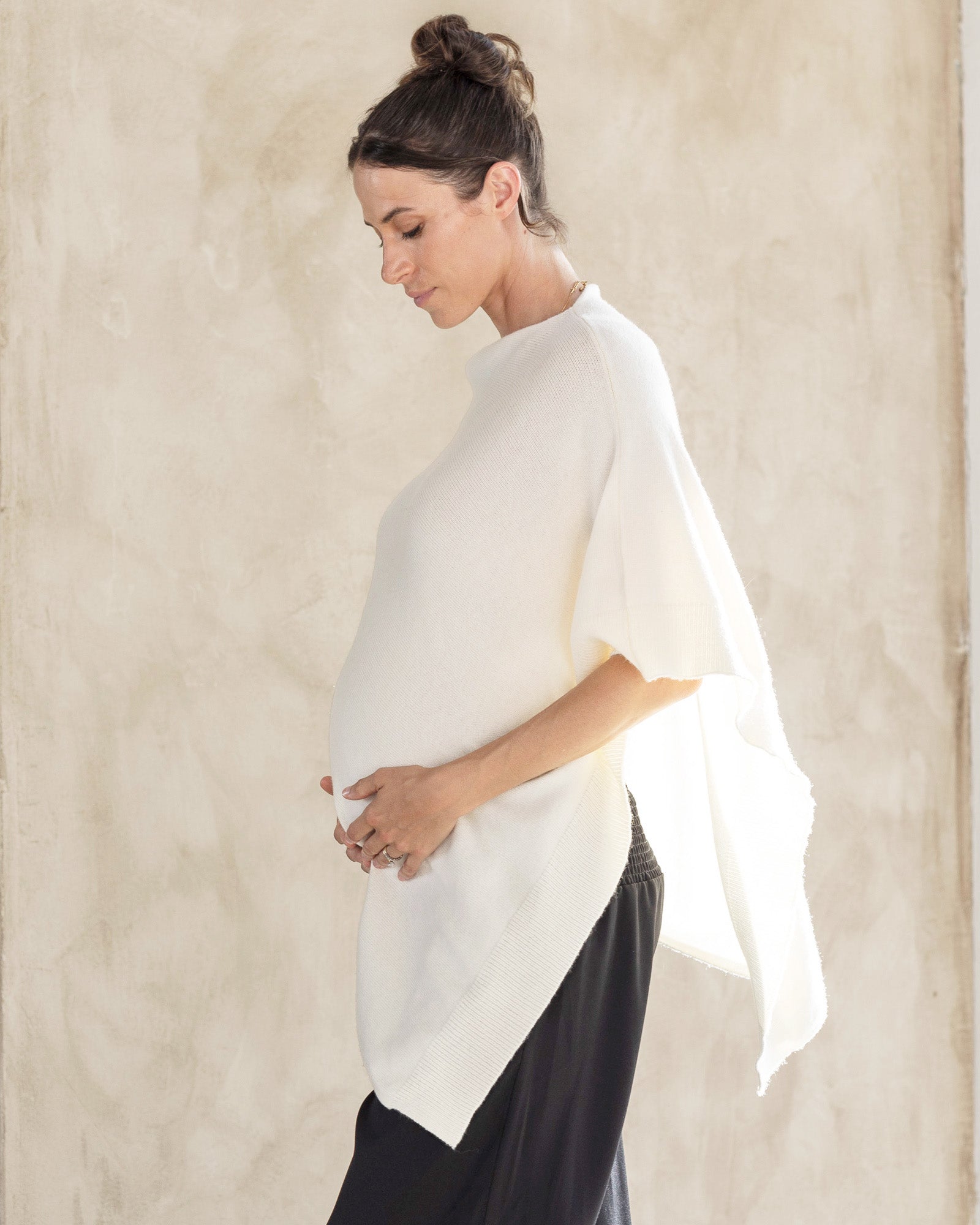 A woman wearing a white maternity nursing poncho over a black outfit, standing against a textured background