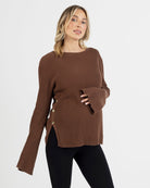 A woman wearing a brown oversized knit maternity jumper with side button opening, long bell sleeves, and a round neckline.