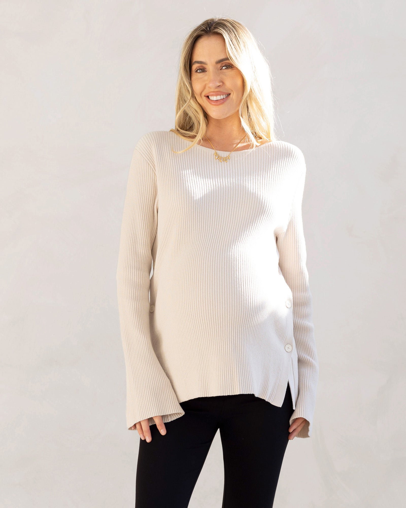 A woman wearing a cream-colored oversized maternity and nursing jumper with long bell sleeves and side button opening for breastfeeding.