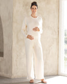 A pregnant woman wearing a cream white 2-piece maternity winter lounge wear set with a long sleeve cropped top and high waist pants.