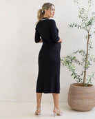 Pregnant Woman wearing a black maternity knit dress standing next to a potted plant against a white wall