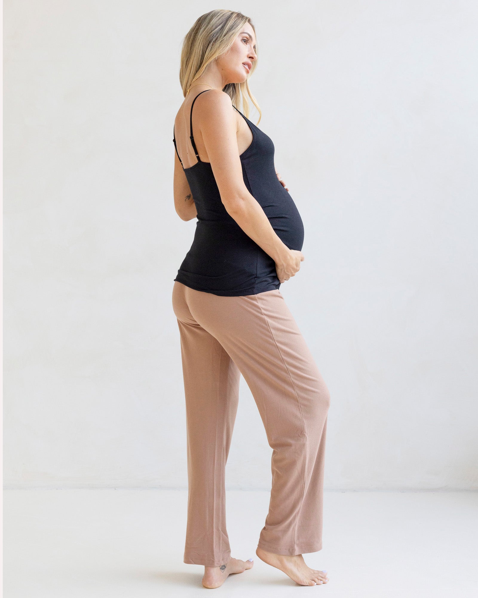 Pregnant woman wearing a black tank top and beige pants on a white background