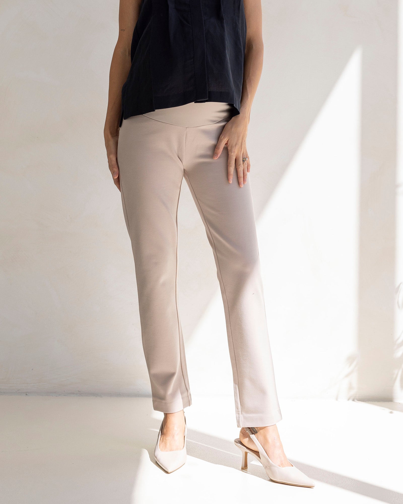 A pregnant woman wearing beige straight cut maternity work pants with a high waist band, paired with a black top and white shoes.