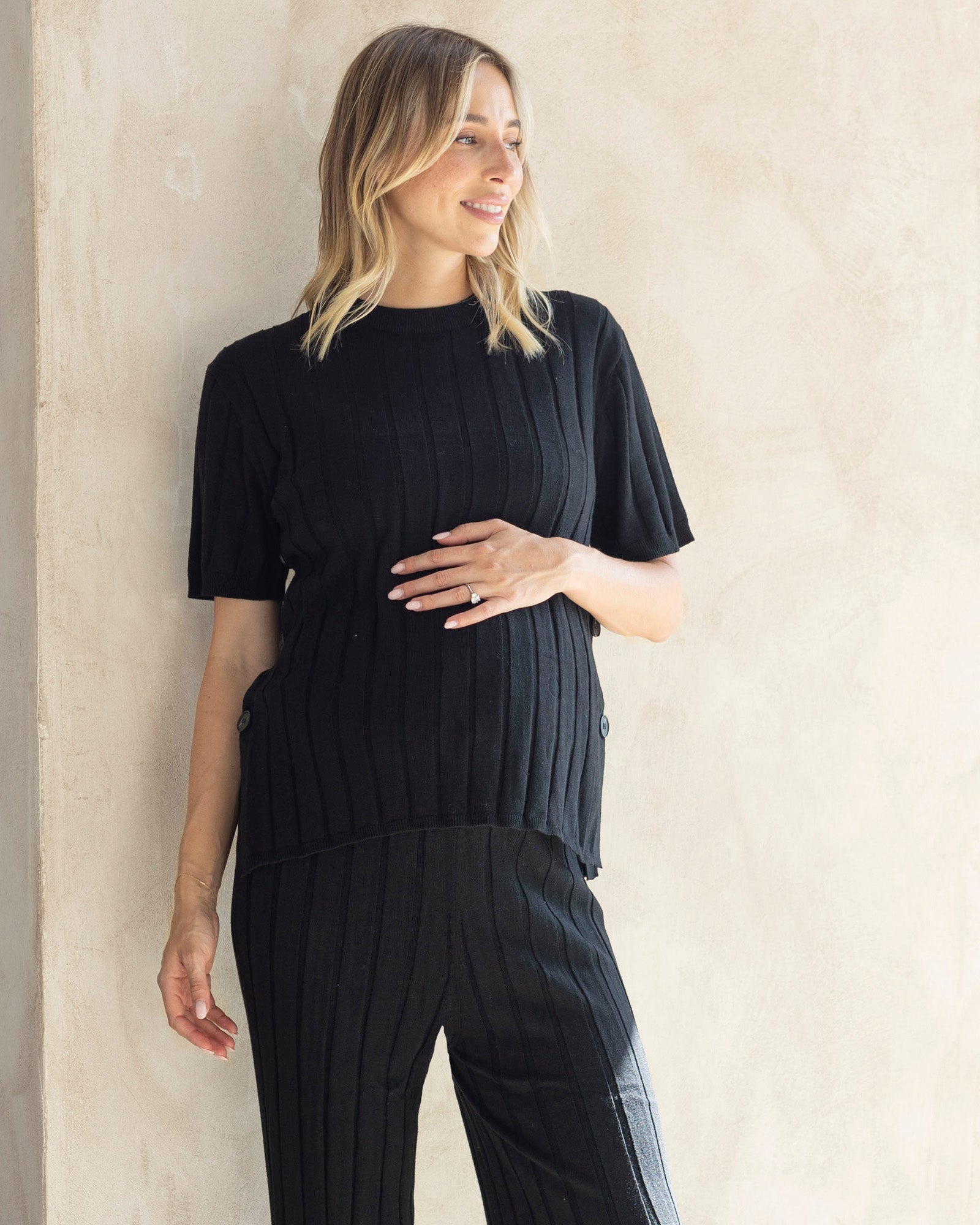 A pregnant woman wearing a black side split maternity top with short sleeves, paired with dark maternity lounge knit pant.