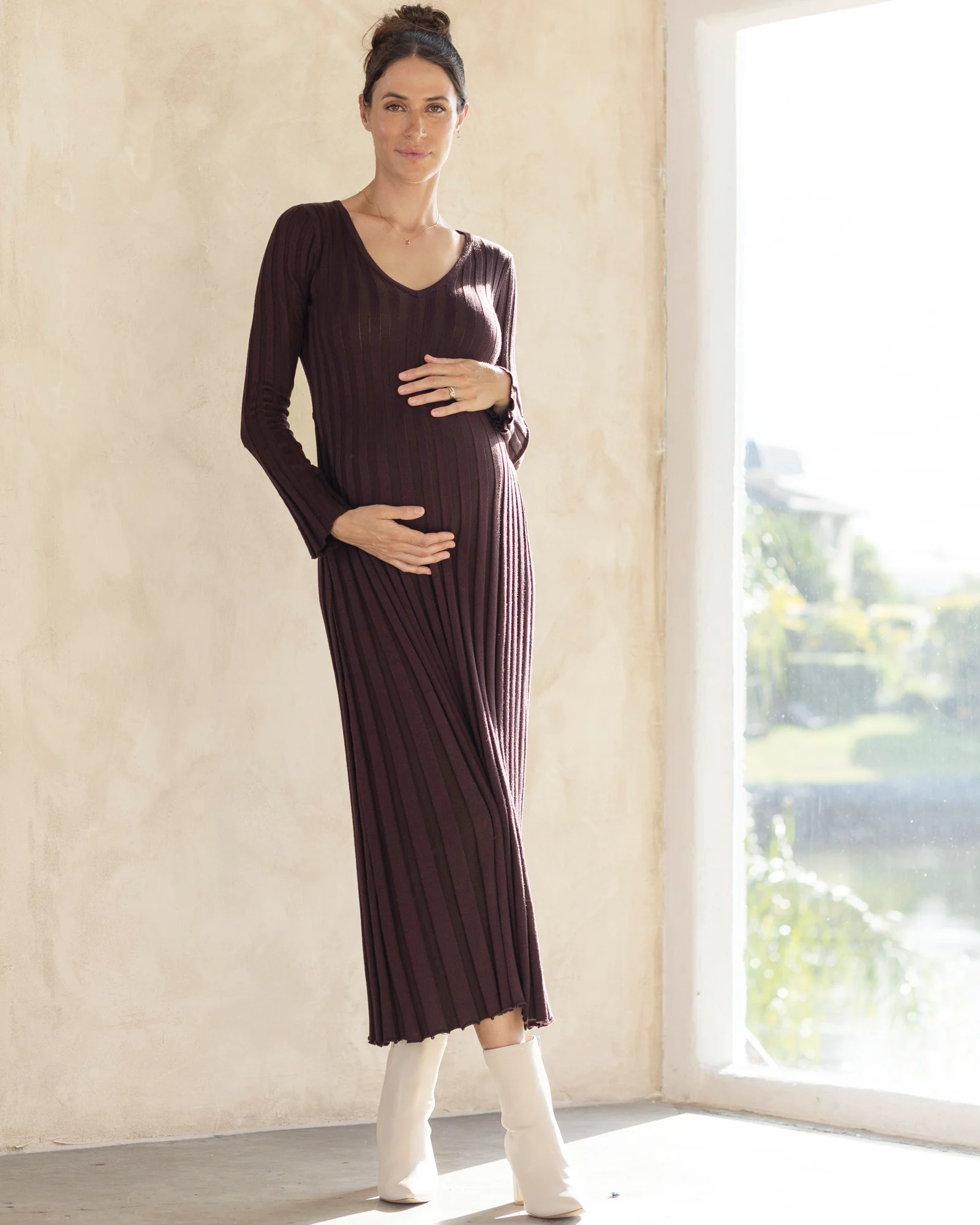 A pregnant woman wearing a dark brown, long-sleeved, knee-length maternity knit bodycon dress with a V-neckline.