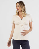 A pregnamt woman wearing a light beige, maternity v-neck work top with short sleeves and a relaxed fit with black maternity Ponti pants
