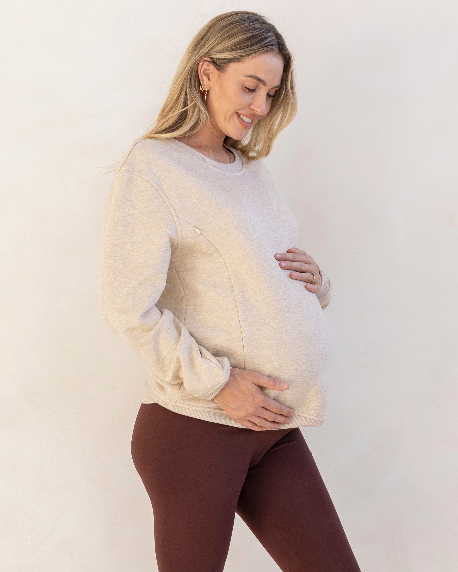 Pregnant woman wearing a light-colored zipper sweater and brown pants against a white background