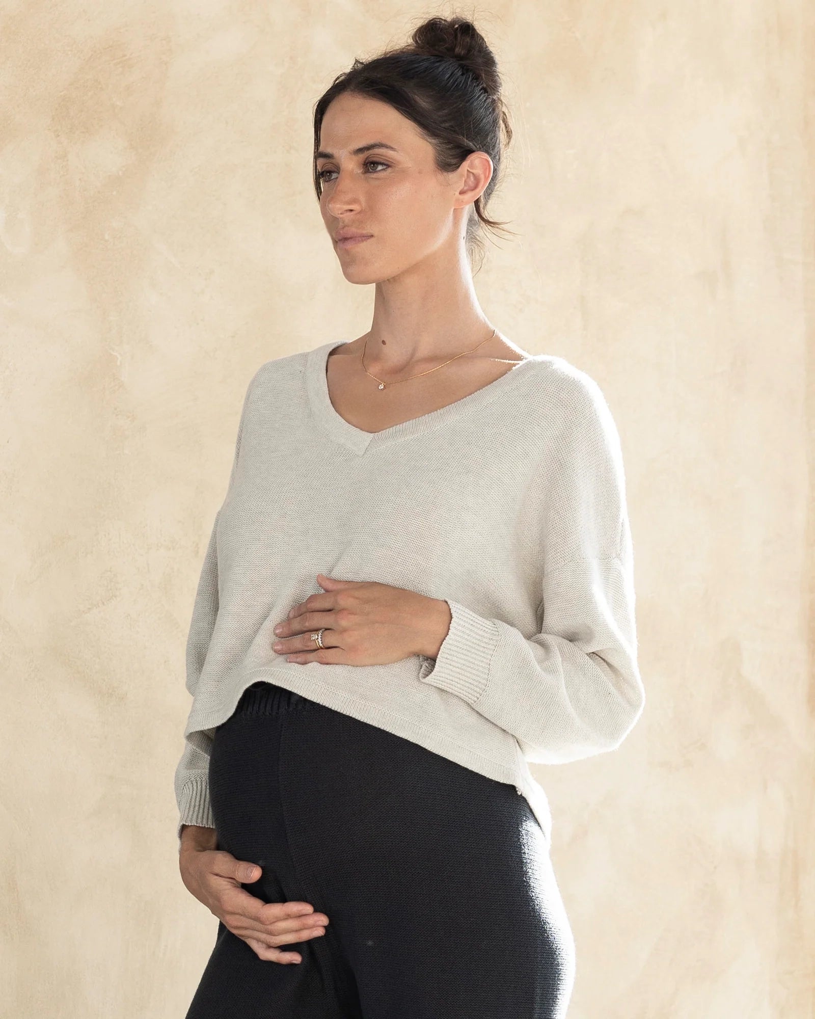 A pregnant woman wearing a white, long-sleeved, V-neckline, cropped length cotton knit maternity sweater and ark grey knitted maternity cotton pants.