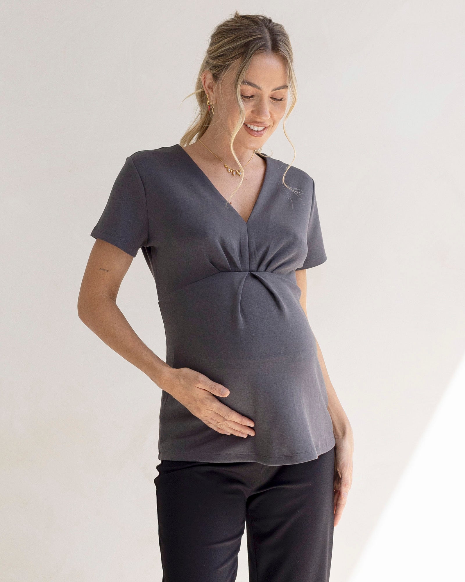 A pregnant woman wearing a slate grey maternity v-neck work top with short sleeves and tucked detailing at the bust.