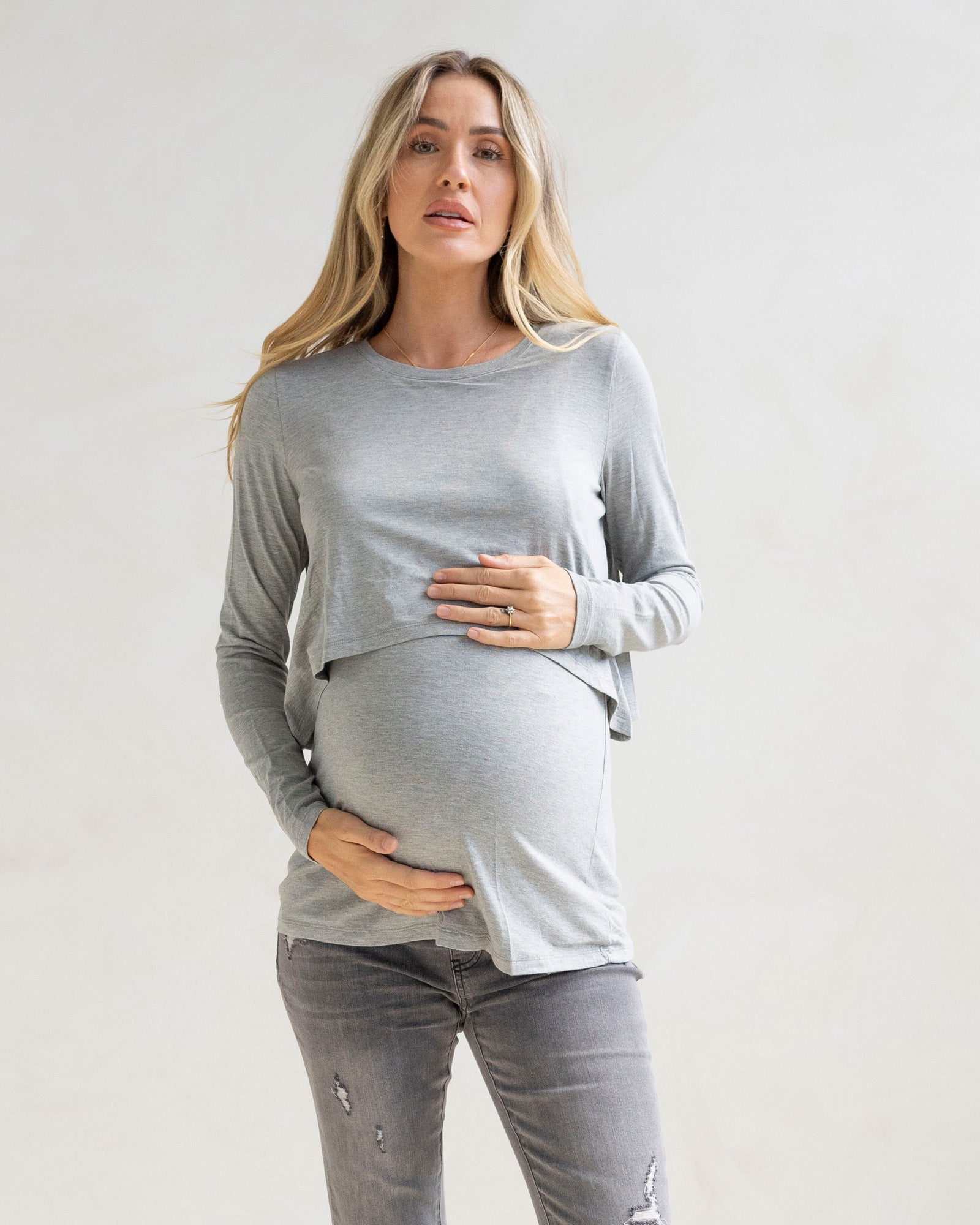 Woman wearing a gray long-sleeve nursing top and gray maternity jean pants against a plain background