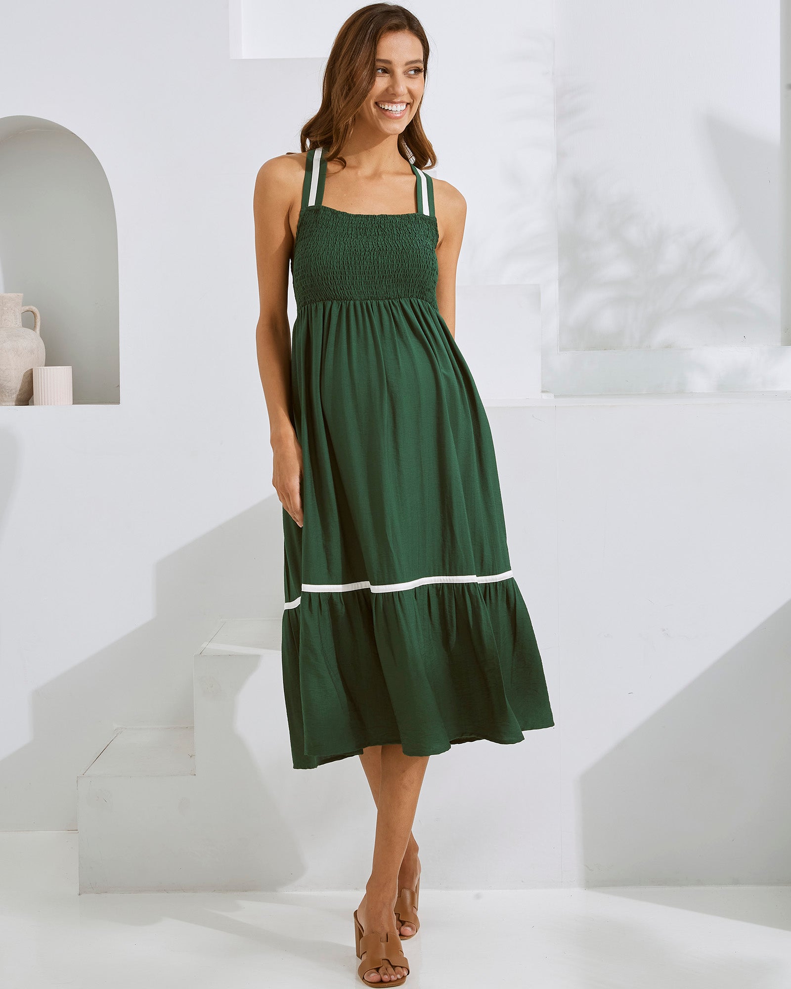 A pregnant woman wearing a green, maternity party dress with contrast ribbon details and adjustable shoulder ties.
