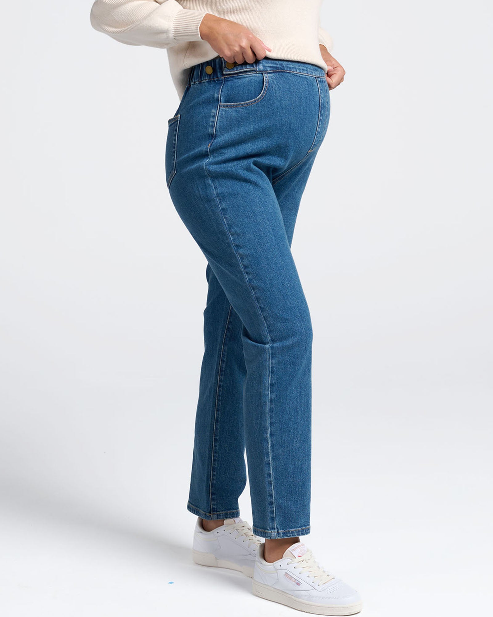 Person wearing high waist maternity blue jeans and white sneakers on a white background