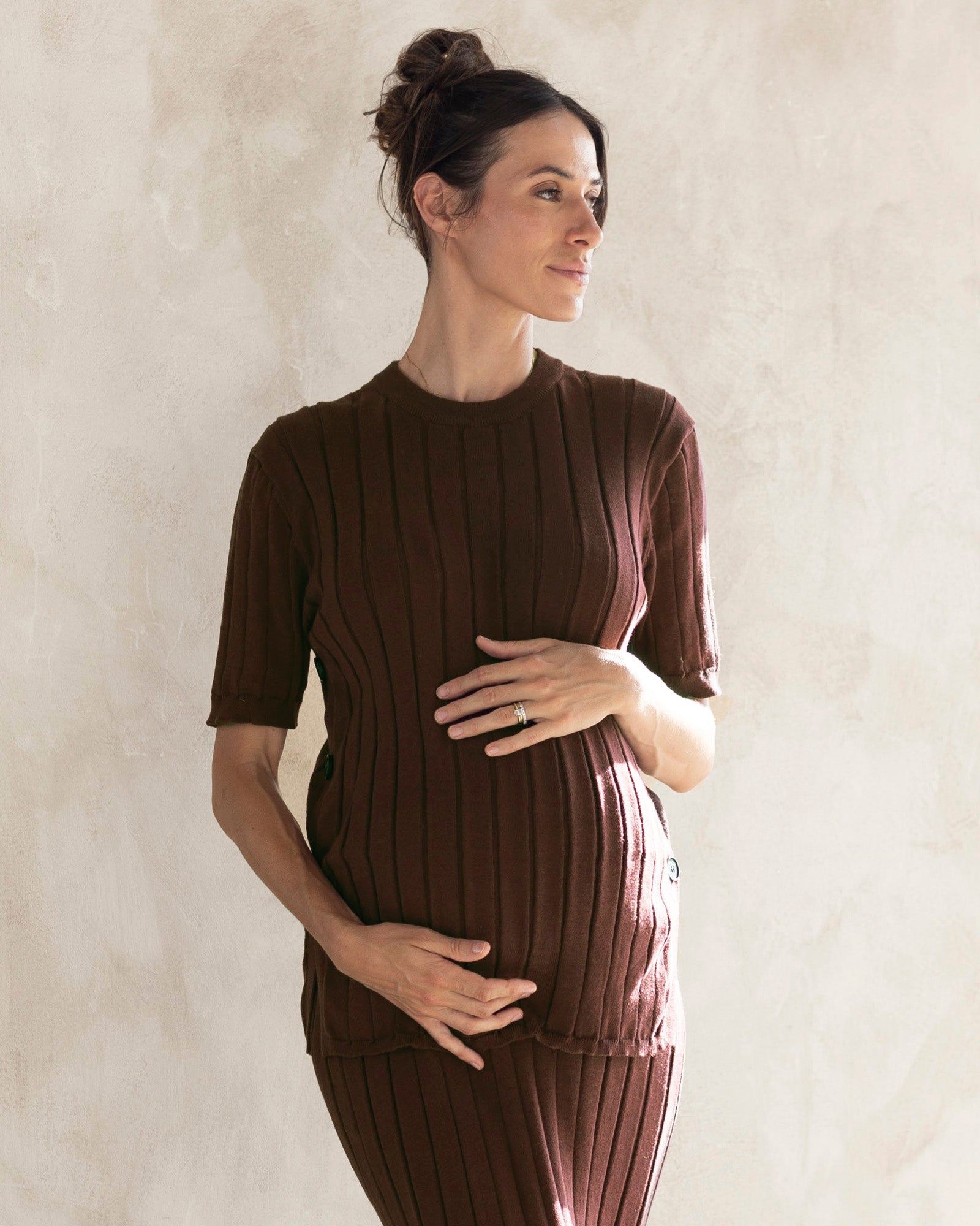 A pregnant woman wearing a brown, side split maternity top with short sleeves and a relaxed fit, made from a cotton blend fabric.