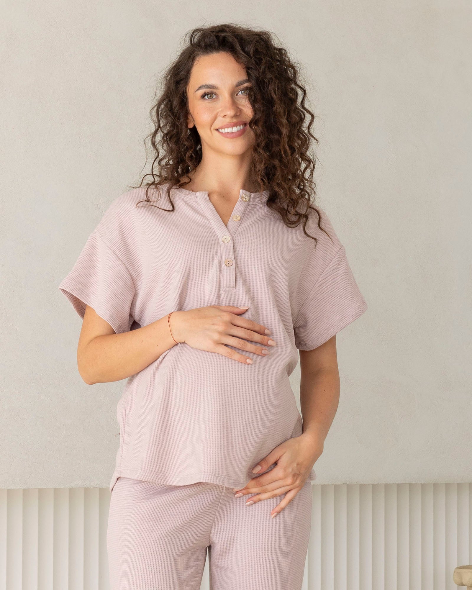 Woman wearing a light pink maternity lounge top and maternity shorts outfit with a plain background