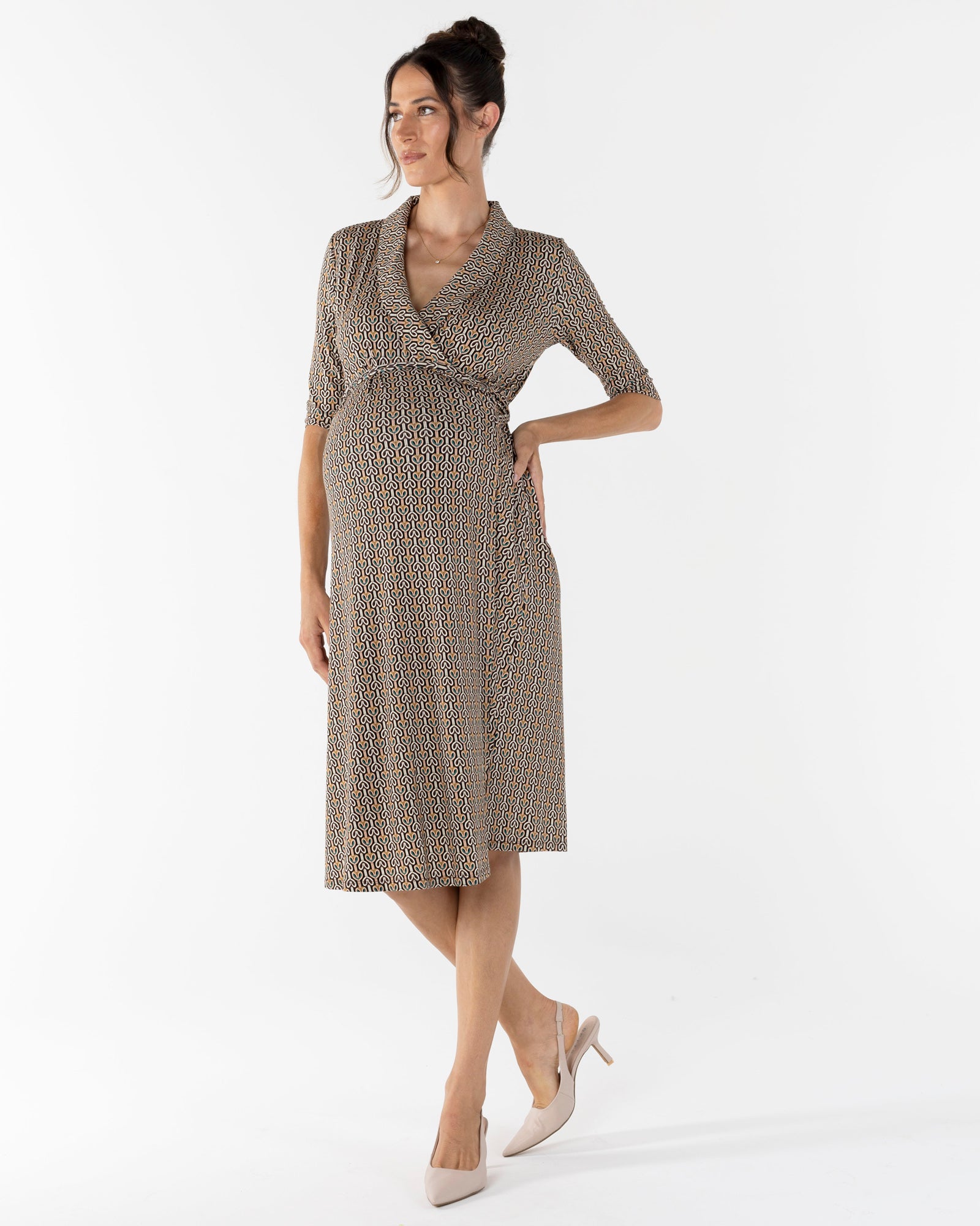 A woman wearing a midi-length maternity wrap dress with a link print. The dress has a wide neckline, short sleeves, and an adjustable tie in a taupe color.