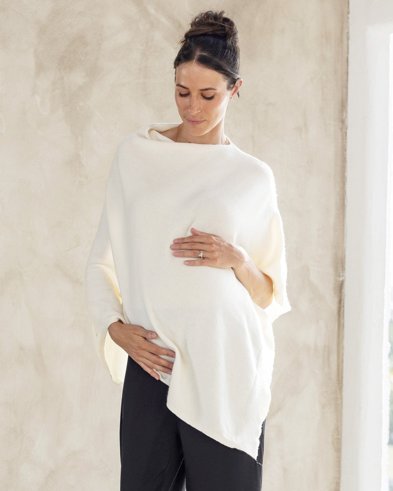 A woman wearing a winter white maternity and nursing poncho with a growing bump, standing indoors.