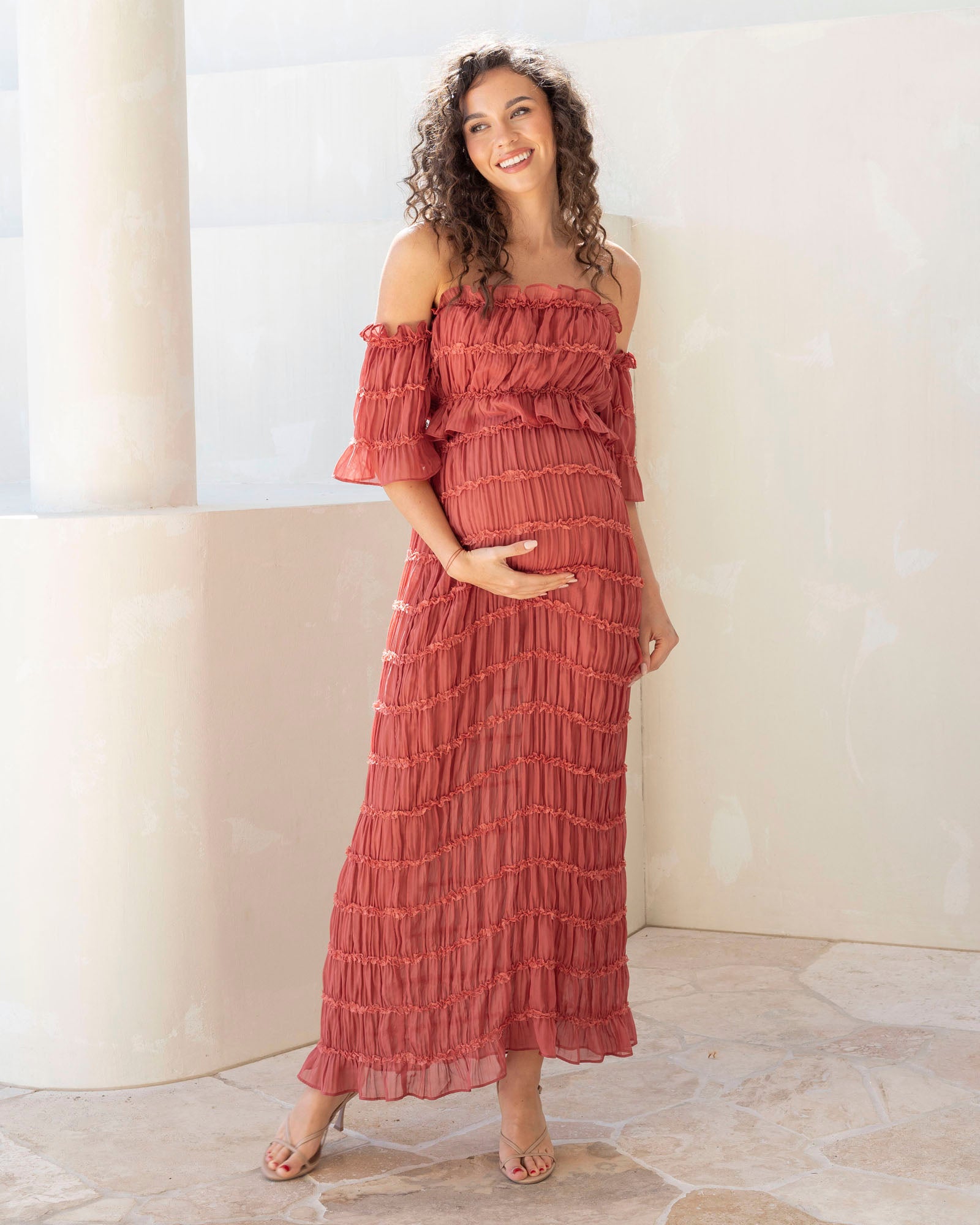 Woman wearing a red off-shoulder maternity baby shower dress standing against a white wall.
