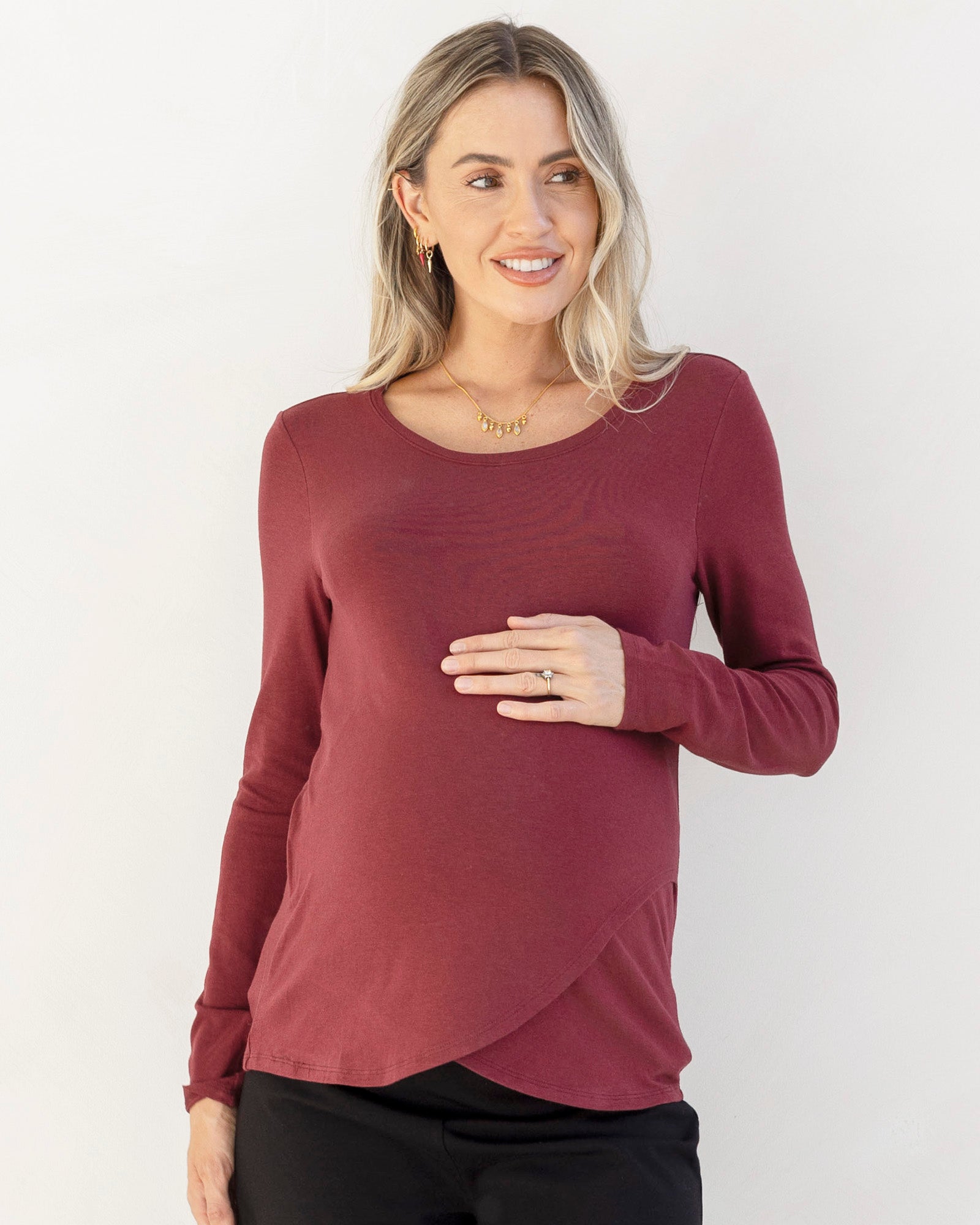 A pregnant woman wearing a long sleeve burgundy nursing top with a round neckline, in a standing pose.