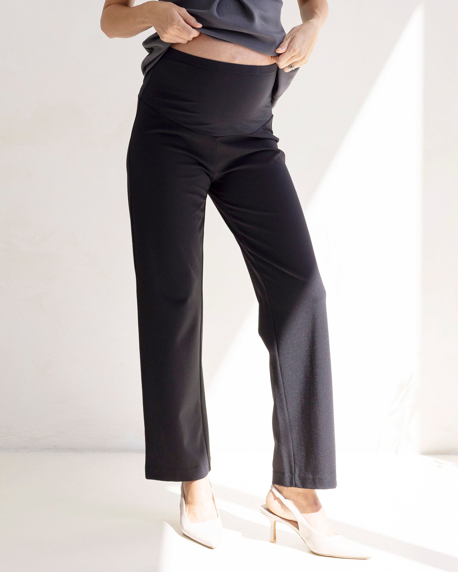 pregnant lady is wearing black maternity work pants and white shoes on a white background