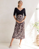 Woman wearing a black maternity dressy top and zebra print skirt standing indoors with a white curtain and plant in the background.