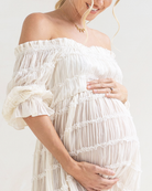 Pregnant woman wearing an off-shoulder white dress against a plain background
