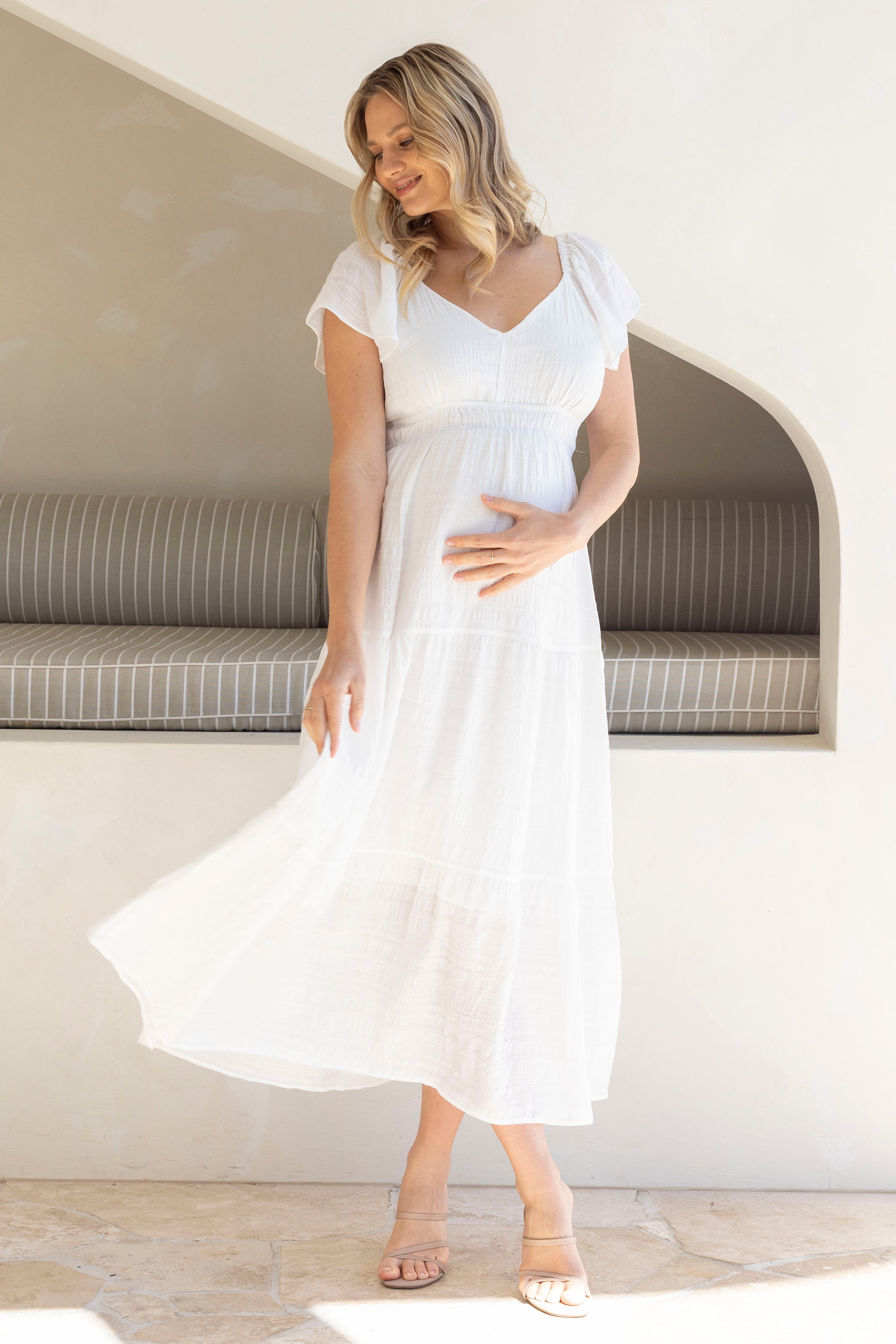 Woman in a white maternity dress standing in a minimalistic room with a bench.