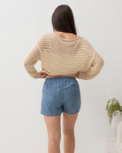 Woman wearing a beige crochet sweater and denim shorts against a plain background