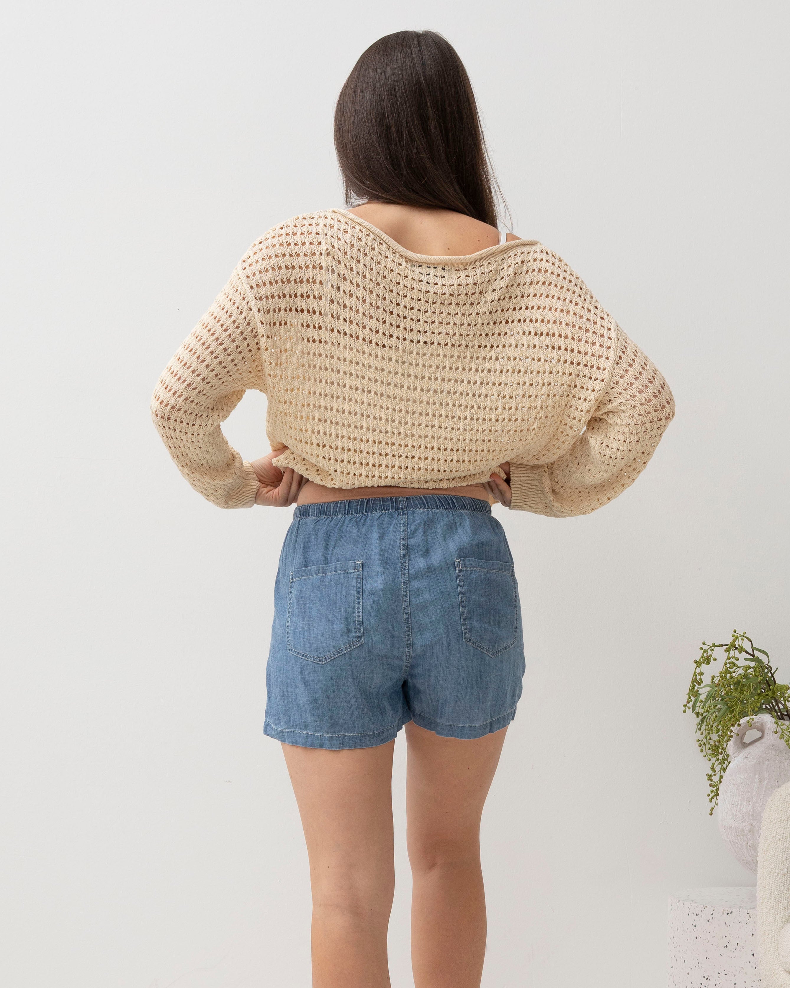 Woman wearing a beige crochet sweater and denim shorts against a plain background