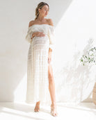 Woman wearing a white off-shoulder maternity photoshoot dress with a slit, standing against a bright background. It is a great baby shower white dress