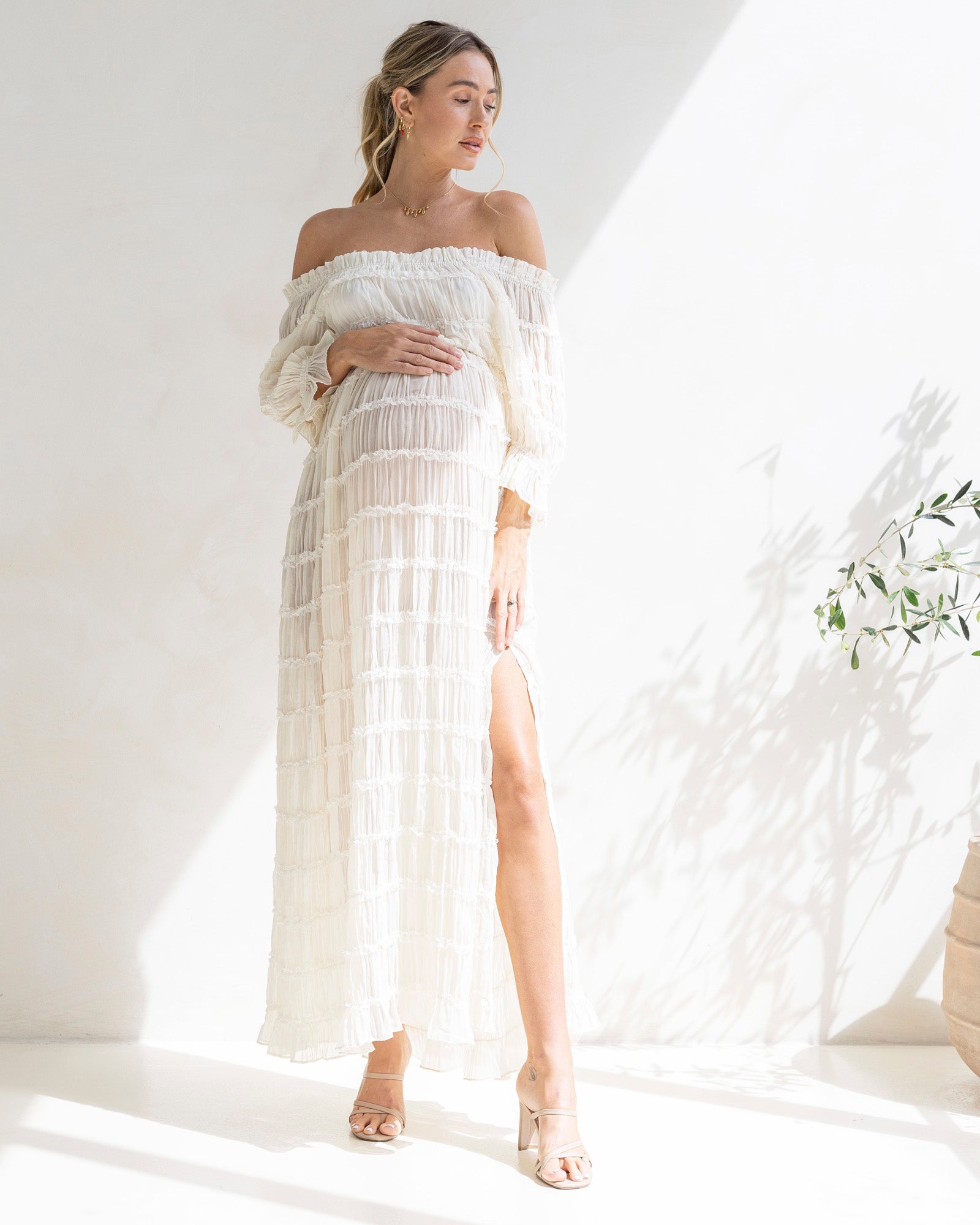 Woman wearing a white off-shoulder maternity photoshoot dress with a slit, standing against a bright background. It is a great baby shower white dress