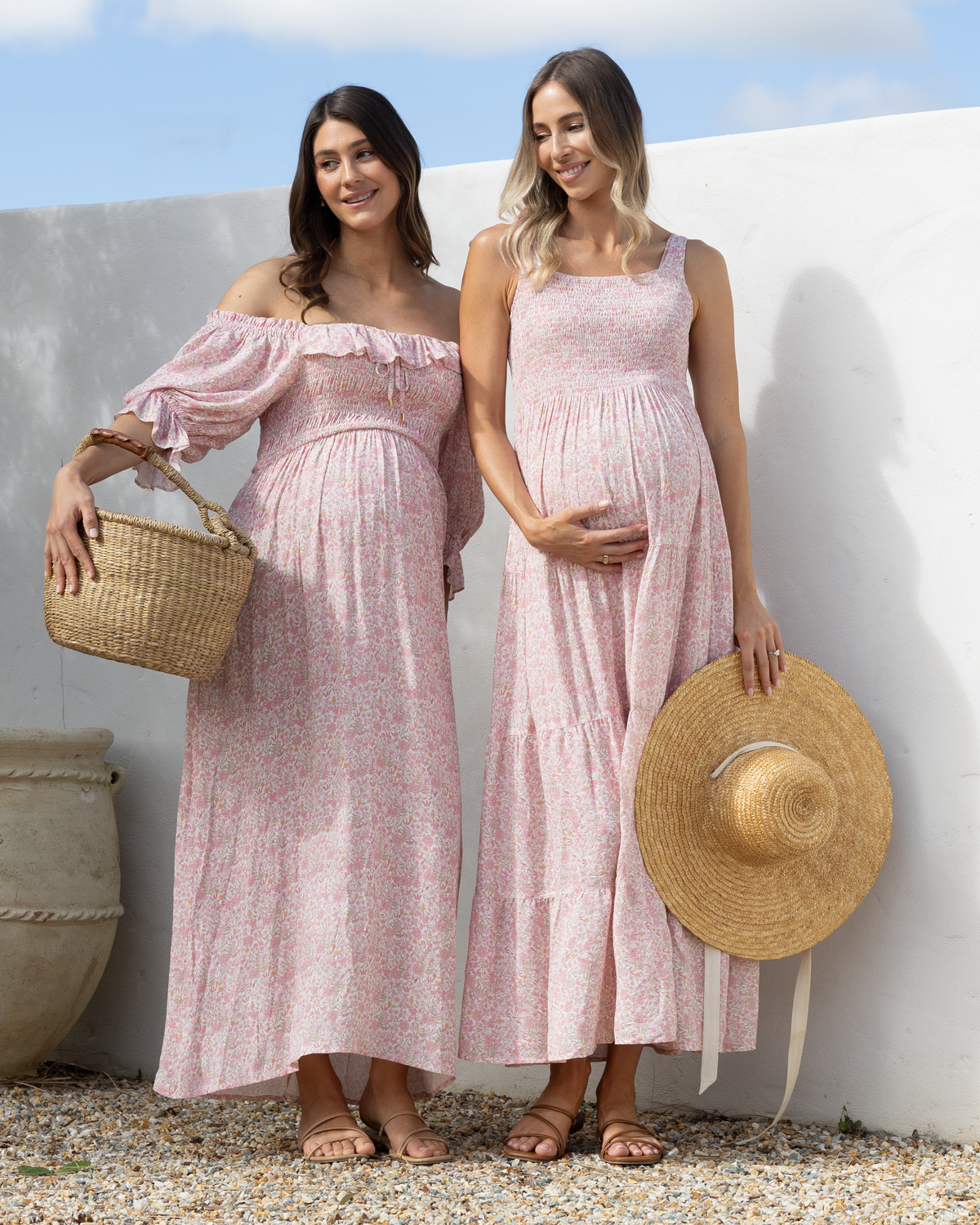 Two women wearing matching light pink floral maxi dresses stand outdoors against a white wall. One dress is off-the-shoulder with puff sleeves, and the other is sleeveless with a scoop neckline.