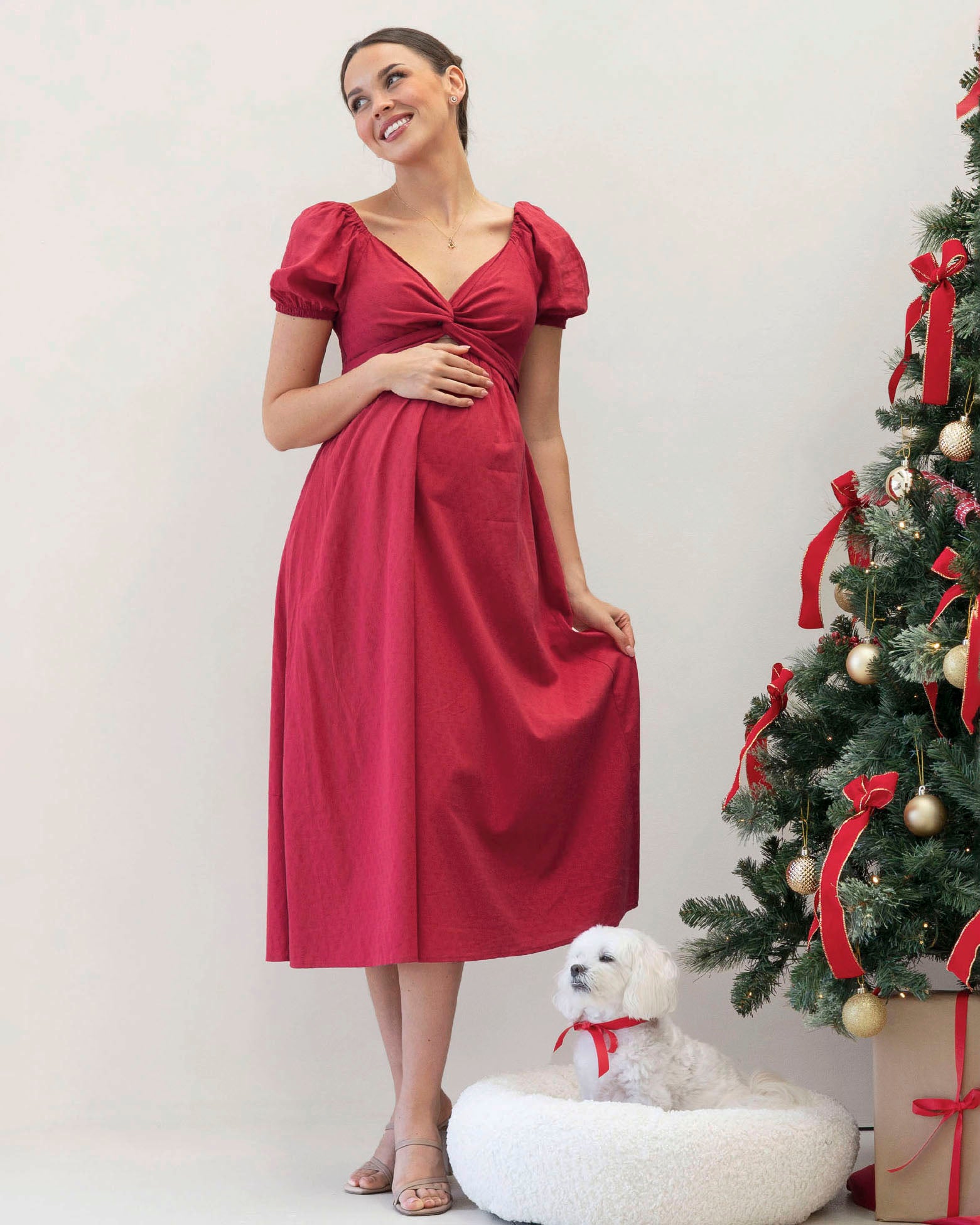 Woman in a red maternity dress standing next to a decorated Christmas tree with a white dog.