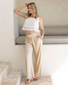 Pregnant lady is wearing a white sleeveless maternity  top and beige maternity linen pants in a minimalistic room