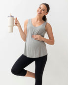 Woman in gray tank top and black leggings holding a white mug with a straw on a light gray background