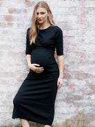 A woman in black angel maternity annabella knit dress against the wall, front (6594399666279)